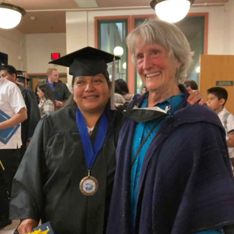This image captures a proud moment at a graduation ceremony. A woman in a black graduation gown and cap, adorned with a blue stole and a medal, is smiling at the camera, standing next to an older woman with short, white hair, who is wearing a blue shirt and a dark blue shawl. They are both smiling and appear to be in a joyous mood. The background is an indoor setting with other people who may be fellow graduates and guests, indicative of the celebratory event.