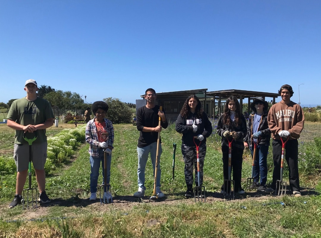 Seven Youthserve volunteers pose with shovels in a field of mown grass, preparing to volunteers for the Homeless Garden project.