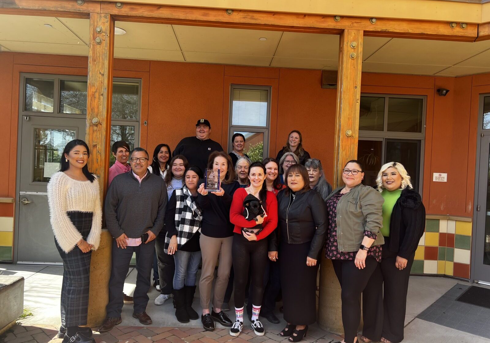 Group photo of 16 diverse volunteers and staff members of the Volunteer Center of Santa Cruz County, standing outside their building. They are smiling and posing together on a sunny day, with one person holding a clear trophy, celebrating their community service achievements.