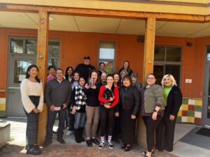 Group photo of 16 diverse volunteers and staff members of the Volunteer Center of Santa Cruz County, standing outside their building. They are smiling and posing together on a sunny day, with one person holding a clear trophy, celebrating their community service achievements.